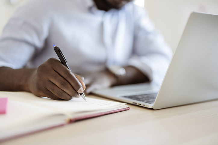 man writing on notepad in front of laptop